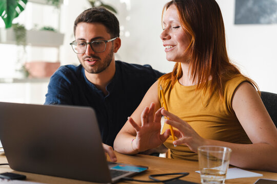A young man and woman sit together at a desk, collaborating on a digital project on their laptop. They focus on the screen while sharing ideas in an office space.