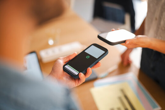 Two coworkers use their smartphones to exchange data or process a digital payment in a modern office. The focus is on the proximity of the mobile devices.