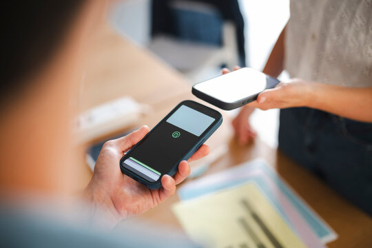 Two people perform a contactless payment transaction by holding their mobile phones near each other in a bright office environment. Digital technology enables modern exchange.