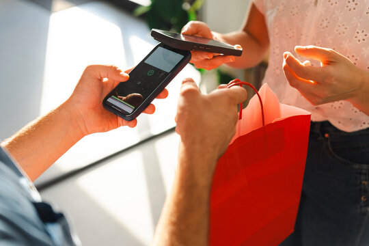 A customer makes a contactless payment by tapping their smartphone against the shopkeeper device. The merchant holds a red paper bag ready for the store purchase.
