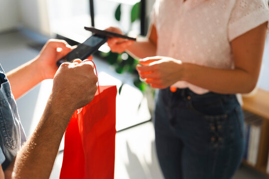 A delivery person hands over a red shopping bag to a female customer. The customer uses her smartphone to confirm the receipt of the package in a bright indoor setting.
