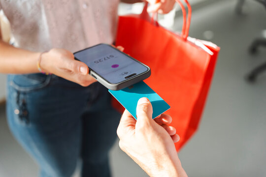 A customer makes a payment by tapping their blue debit card against a merchant's mobile phone device. The shopper carries a red bag after completing a store purchase.