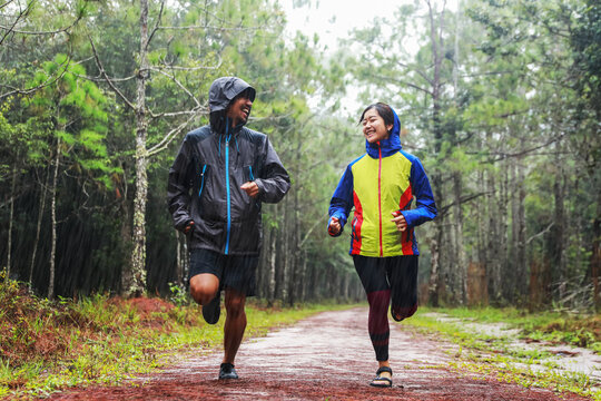 Happy Asian couple wearing waterproof jackets jogging together in rainy pine forest path during outdoor travel adventure vacation