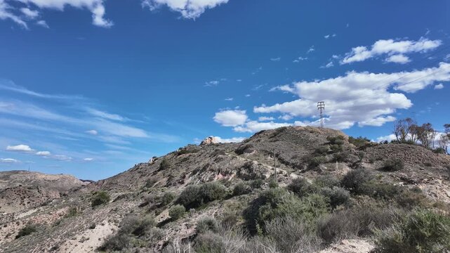 A timelapse shows clouds drifting over a dry rugged hillside with sparse vegetation and a power line, ideal for nature and landscape documentaries