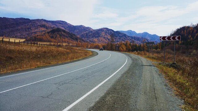 Silver SUV drives along curving mountain road. Autumn colors surround winding asphalt path. Road sign warns of sharp turn ahead. Vehicles move through scenic rural landscape