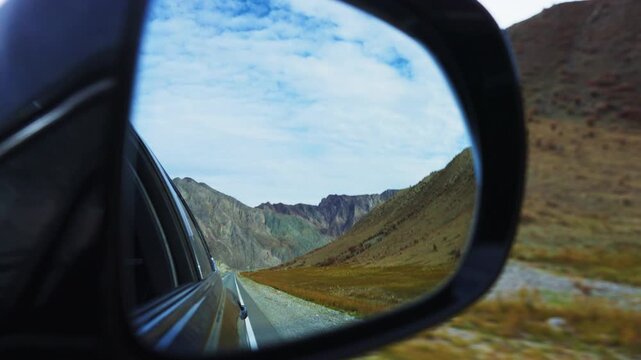 View through car side mirror shows winding mountain road. Mountains stretch under blue sky with clouds. Road curves through rocky terrain with grassy slopes. Scene evokes freedom, adventure