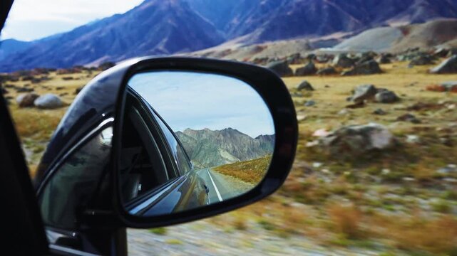 Car side mirror reflects winding mountain road. Mountains rise sharply against blue sky. Grassland stretches beside rocky terrain. Motion blur shows fast travel through scenic route