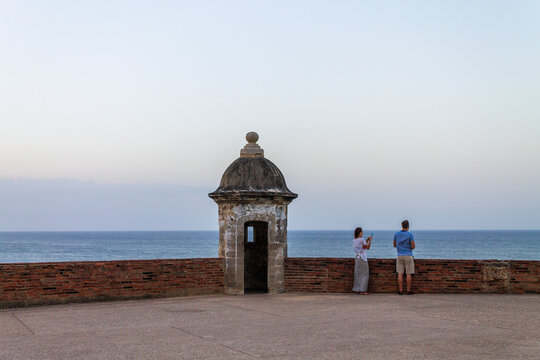 San Juan, Puerto Rico - 22 March 2014: View of a historic stone sentry box at Castillo San Felipe del Morro with two people standing by the brick wall overlooking the ocean.