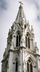 Fototapeta premium Church steeple with elaborate stonework rises against backdrop of cloudy sky, white facade adorned with arched stained glass windows, intricate lacing, prominent cross crowns pinnacle.