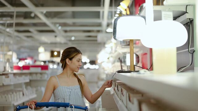 Happy young Asian woman pushing a shopping cart while carefully choosing table lamps in a large retail furniture store. She checks the price tag to buy new lighting for her modern home interior.