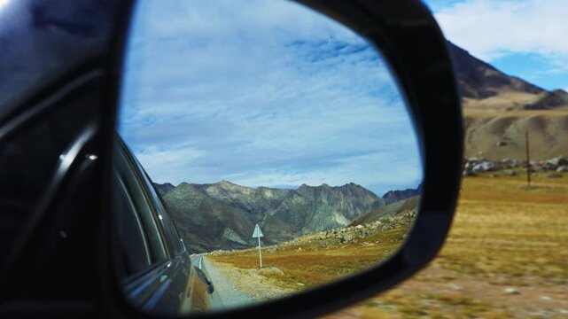 Car side mirror reflects mountain road under blue sky. Journey through scenic highlands with winding road. Serene view evokes freedom and adventure. Ideal for travel, road trip