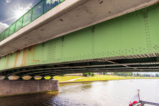 The girder of a riveted steel plate-girder bridge spanning a calm body of water. Bright sunlight shining through the railing. Green anti-corrosion coating, structural stiffeners, rust bleeding down. 