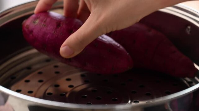 Hand putting raw purple sweet potatoes into a steamer pot to steam, Asian cooking style