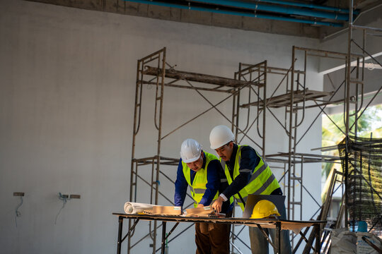 Two Asian construction engineers wearing safety helmets and vests discussing project plans with blueprints, material samples, and a laptop at an indoor construction site.