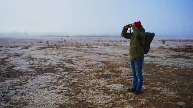 Person stands in frosty field, wearing red hat and green jacket. They raise hands to cover ears from cold wind. Ground covered in thin ice and dry grass. Distant horizon shrouded in mist, giving quiet