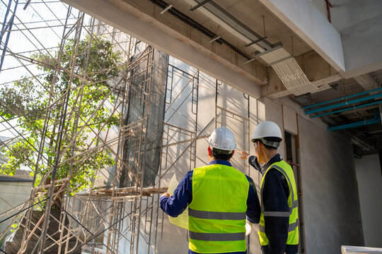 Rear view of two Asian construction engineers in safety gear inspecting the building site, holding blueprints and pointing at scaffolding.