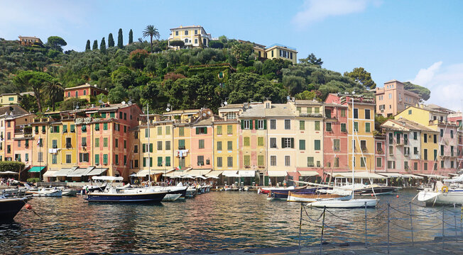 Portofino, Italy - 30 August 2018: View of the colorful historic buildings along the harbor in Portofino, with yachts moored in the water and lush green hills in the background.