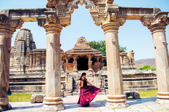 Attractive Indian woman joyfully dancing and twirling between four intricately carved stone pillars at Sahastrabahu Temple, Nagda, Rajasthan, with a beautiful heritage and scenic backdrop.