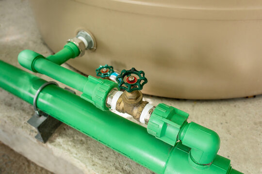 Close-up of green valve wheel mounted on pipe fitting near beige water storage tank, plumbing infrastructure detail