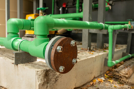 Green PVC pipes connected with rusty flange joint mounted on concrete base near building, plumbing infrastructure detail