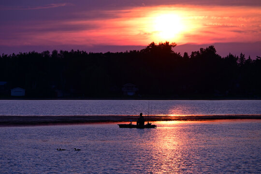 A lone fishermen sitting on a platform boat fishing boat as the sun sets behind him