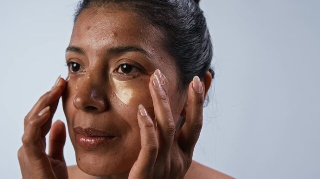 Young Hispanic woman applying golden hydrogel eye patches under eyes, smoothing them with fingers and smiling. Clip is ideal for skincare product commercials and beauty marketing campaigns