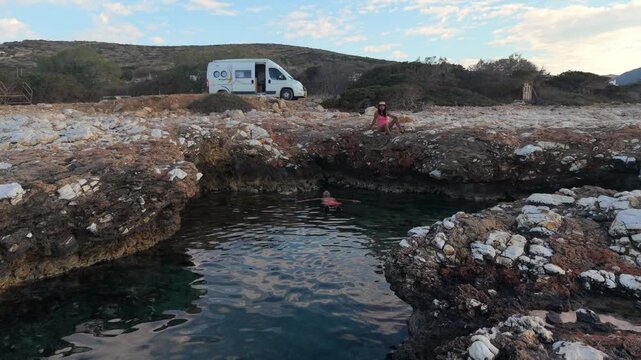 Explore the tranquil landscape of Paros Island in Greece, where beautiful rocky shores meet crystal-clear waters. A couple relaxes by the ocean with a camper van motorhome in the background