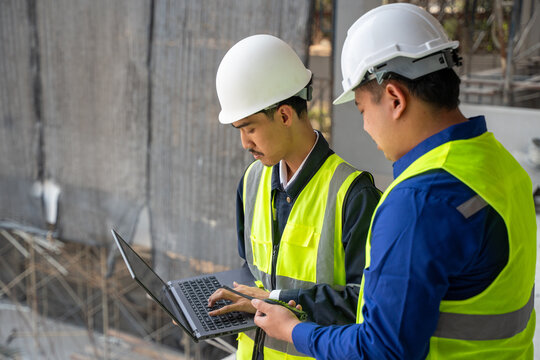 Two Asian construction engineers in safety helmets and reflective vests using a laptop and holding a walkie-talkie at an industrial building site.