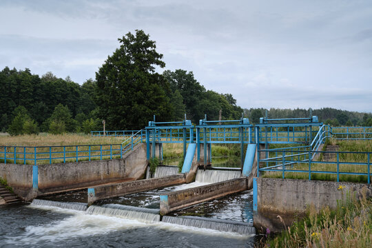 Wide view of a multi-gate weir with blue sluice gates and water flowing through concrete channels, illustrating river engineering and water management.