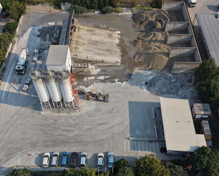 Aerial view of a concrete plant in an industrial zone with cement silos, aggregate piles, a front loader, a concrete mixer truck, and parked cars in Belgrade, City of Belgrade, Serbia.
