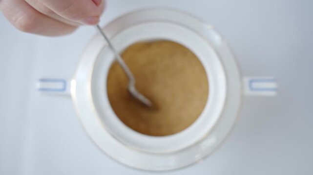 A teaspoon of brown sugar is lifted from a white sugar bowl and then poured back in slow motion.