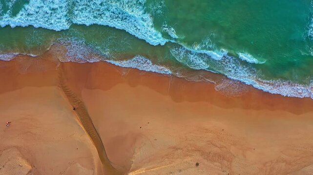 aerial top view of ocean waves gently crashing into a sandy beach, creating a natural contrast between deep green water and warm golden sand. The scene highlights coastal textures and seaside beauty.