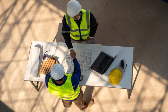 Top view of two Asian construction engineers shaking hands over a table with blueprints, a laptop, and material samples at a building site.