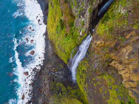 Aerial view of a waterfall cascading down a steep, mossy, rocky cliff into the Atlantic Ocean, with waves crashing against the dark pebble beach below in Madeira, Portugal.