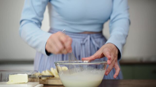 Woman whisks cream in glass bowl in cozy kitchen, close-up, 25 fps