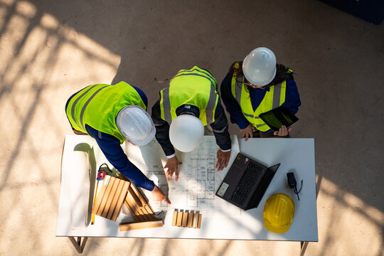 Top view of a team of three Asian construction engineers in safety gear discussing building plans over blueprints, a laptop, and a tablet at a construction site.