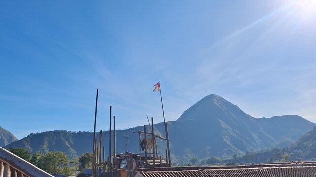 The Indonesian red and white national flag (Merah Putih) flying on a traditional bamboo pole under a clear blue sky, with a beautiful green mountain and rural rooftop in the background.