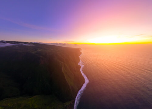 Aerial view of the dramatic coastline of Madeira at sunset, with steep cliffs meeting the Atlantic Ocean under a vibrant purple and orange sky, Madeira, Portugal.