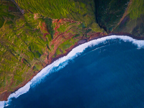Aerial view of the dramatic coastline of Madeira with lush green cliffs, striking red rock formations, and deep blue ocean waves crashing against the shore in Madeira, Portugal.
