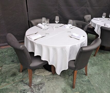 Mesa redonda con mantel blanco y sillas tapizadas con patas de madera en un restaurante o sal&oacute;n de hotel, con cubiertos, servilletas y copas de vino sobre la mesa.