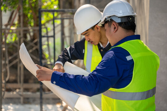 Two Asian construction engineers in safety helmets and vests closely examining and discussing a blueprint at an outdoor building site.