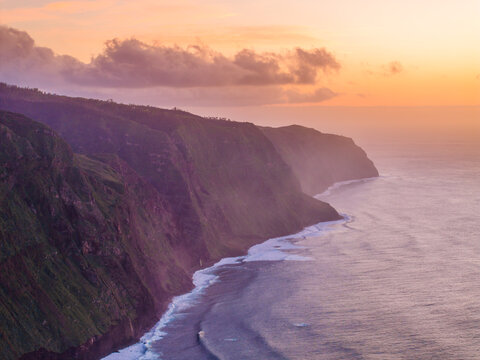 Aerial view of the dramatic coastal cliffs of Madeira at sunset with golden light hitting the rugged landscape and waves crashing against the shore in Madeira, Portugal.