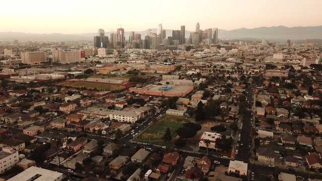 Aerial view of the Los Angeles skyline with urban residential neighborhoods, a soccer field, and distant mountains under a soft sunset sky in Los Angeles, California, United States.