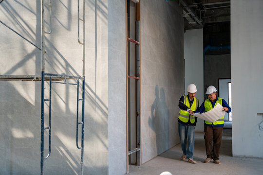 Two Asian construction engineers in safety gear walking and discussing a blueprint inside an unfinished building with scaffolding shadows.