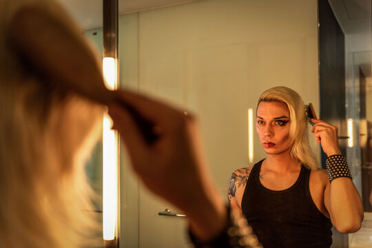 Person with long hair and tattoos applies makeup in front of a mirror.