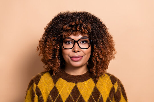 Young woman with curly hair and glasses wearing a yellow argyle sweater, smiling confidently in a studio portrait