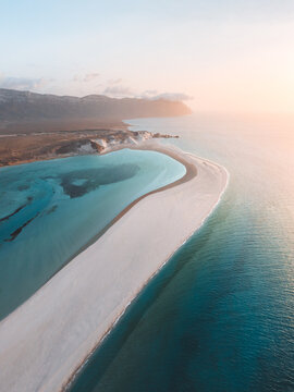 Aerial view of Detwah Lagoon Ramsar Site featuring a pristine white sandbar, turquoise waters, and arid coastal terrain under soft golden hour light in Socotra, Yemen.