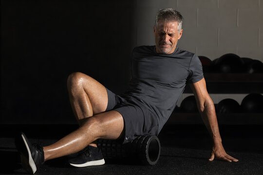 Mature man foam rolling his leg on a gym floor, grimacing with an expression of discomfort and pain during muscle recovery.