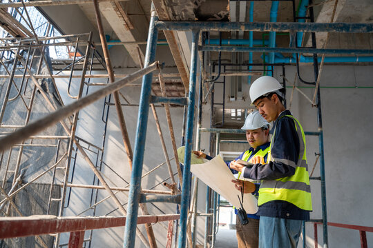 Two Asian construction engineers in safety gear holding a walkie-talkie and discussing a blueprint amidst scaffolding at an indoor building site.