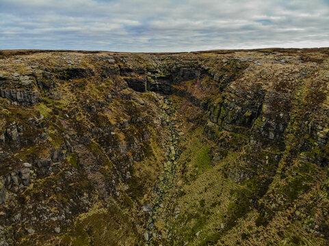 Aerial view of the rocky gorge at High Peak with steep mossy cliffs, scattered boulders, and a narrow stream bed under a cloudy sky in High Peak, England, United Kingdom.